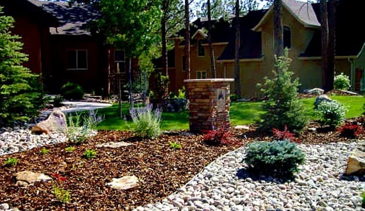 Landscaped front yard with a stacked-stone pillar, river rock, wood mulch, blue spruce and lavender in front of two stucco homes